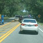 Traffic backs up along a two-lane road leading to the Rock the Country festival, with vehicles including a white Toyota sedan and a truck towing equipment stopped in a line. Law enforcement vehicles with flashing lights and cones are visible ahead, managing entry and flow. Trees with hanging moss arch over the shaded roadway, and a yellow traffic sign warns of a side road ahead.