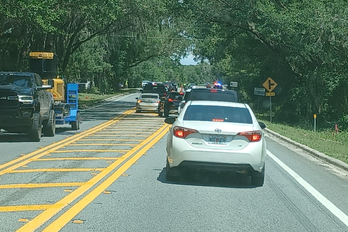 Traffic backs up along a two-lane road leading to the Rock the Country festival, with vehicles including a white Toyota sedan and a truck towing equipment stopped in a line. Law enforcement vehicles with flashing lights and cones are visible ahead, managing entry and flow. Trees with hanging moss arch over the shaded roadway, and a yellow traffic sign warns of a side road ahead.