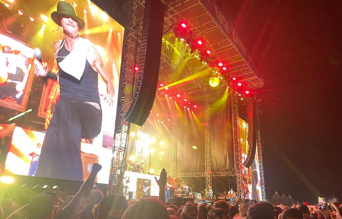 A performer energizes the crowd during a nighttime set at Rock the Country, with fans cheering under bright stage lights and a massive video screen.