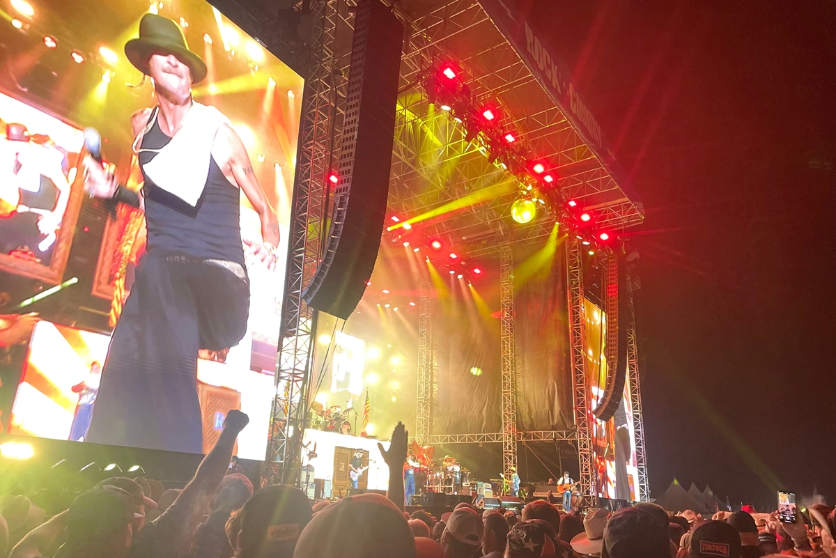 A performer energizes the crowd during a nighttime set at Rock the Country, with fans cheering under bright stage lights and a massive video screen.