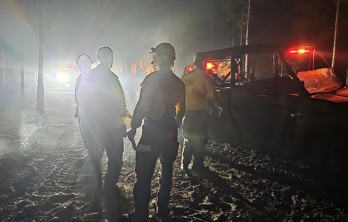 Silhouetted firefighters stand near utility vehicles amid thick smoke and bright lights during nighttime wildfire response in the Ocala National Forest.