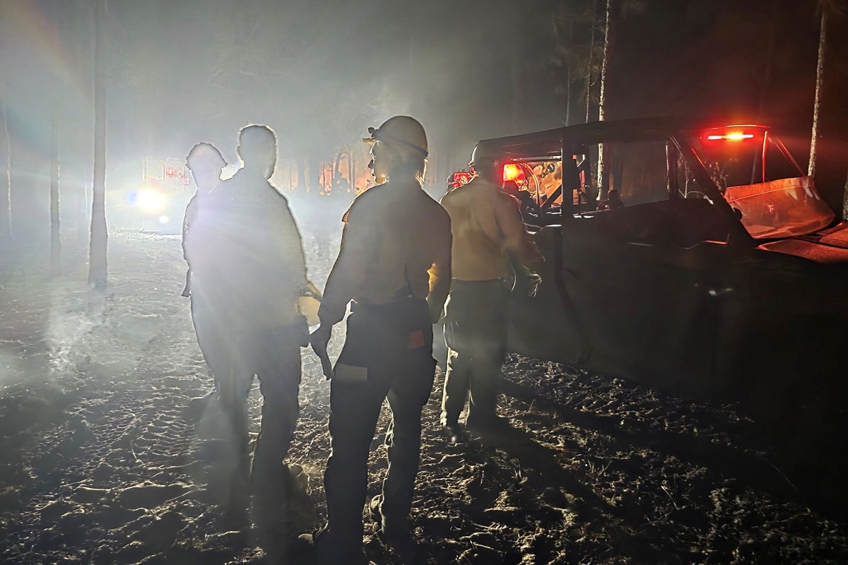 Silhouetted firefighters stand near utility vehicles amid thick smoke and bright lights during nighttime wildfire response in the Ocala National Forest.