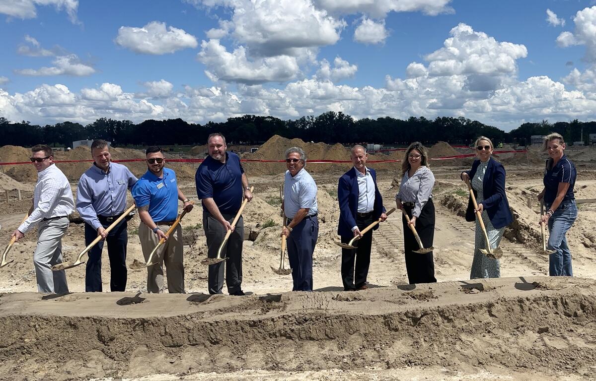 A group of nine people holding shovels pose for a photo during a groundbreaking ceremony at a construction site under a sunny sky, marking the start of a new Jenkins Automotive Volvo/Subaru dealership project in Ocala.