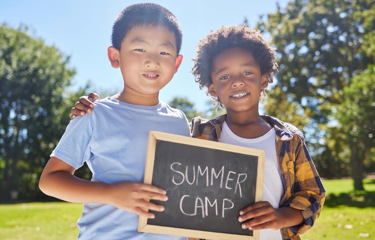Two young children holding a chalkboard that says “Summer Camp” while standing outside on a sunny day.
