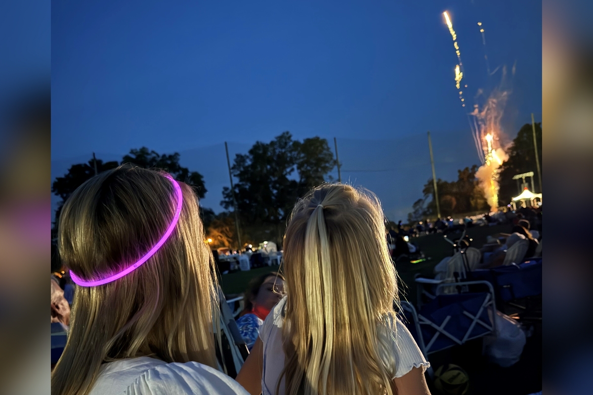 A woman wearing a glowing purple headband and a young girl with blonde hair watch fireworks light up the night sky during an outdoor concert at the Ocala Golf Club.