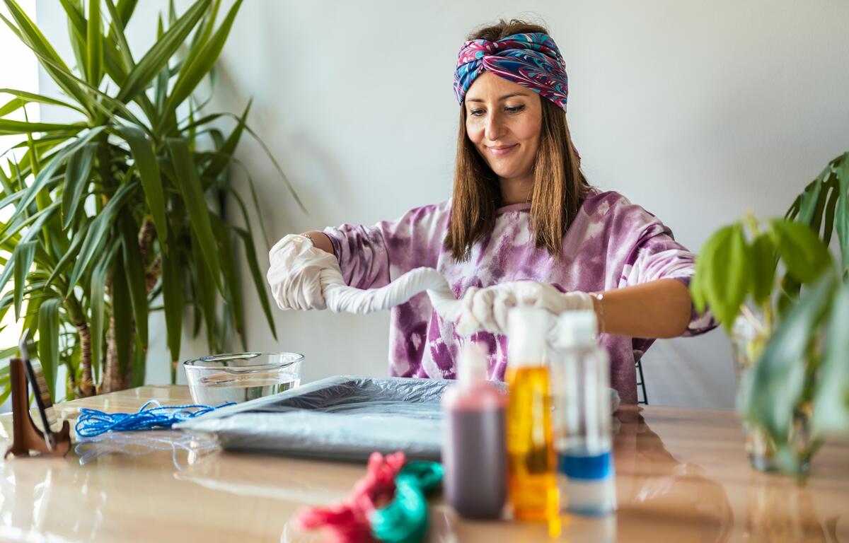 Woman wearing gloves and a colorful headband creates a tie-dye design at a table with art supplies.