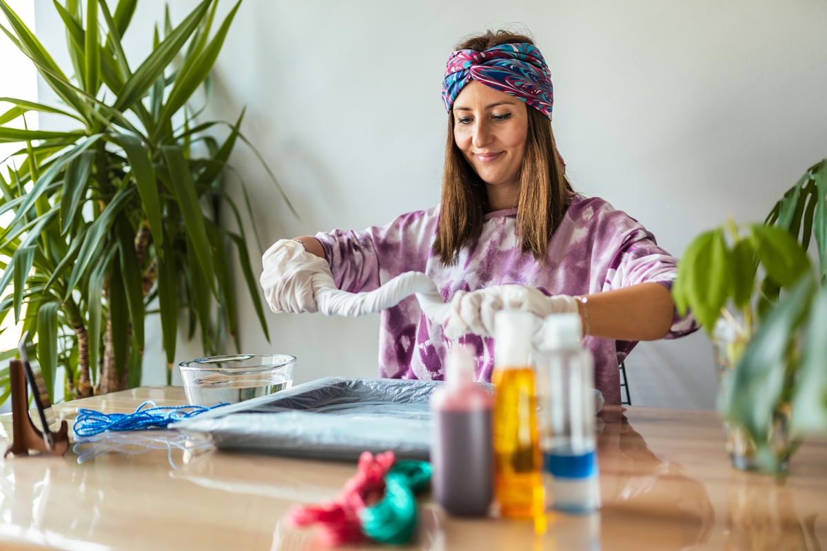 Woman wearing gloves and a colorful headband creates a tie-dye design at a table with art supplies.
