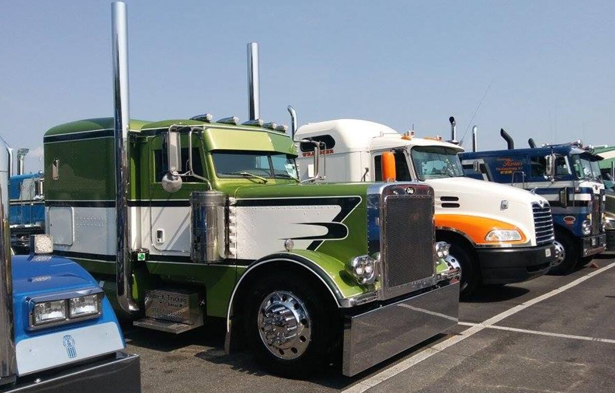 Row of colorful custom semi-trucks parked in a lot, including green, white, and blue designs, displayed for a truck show competition.
