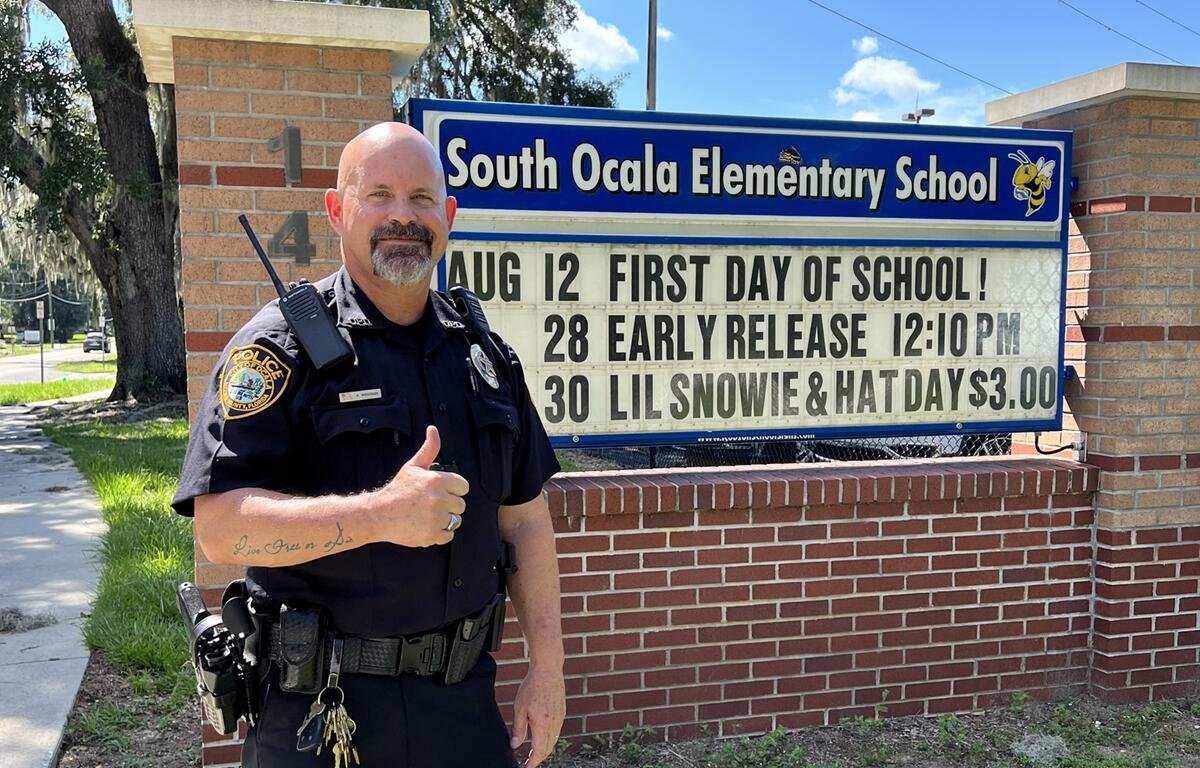 Police officer Roger Wissinger stands smiling with a thumbs-up in front of the South Ocala Elementary School sign, which displays dates for the first day of school, early release, and hat day.
