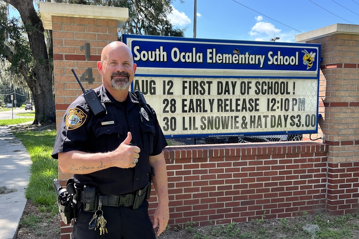 Police officer Roger Wissinger stands smiling with a thumbs-up in front of the South Ocala Elementary School sign, which displays dates for the first day of school, early release, and hat day.