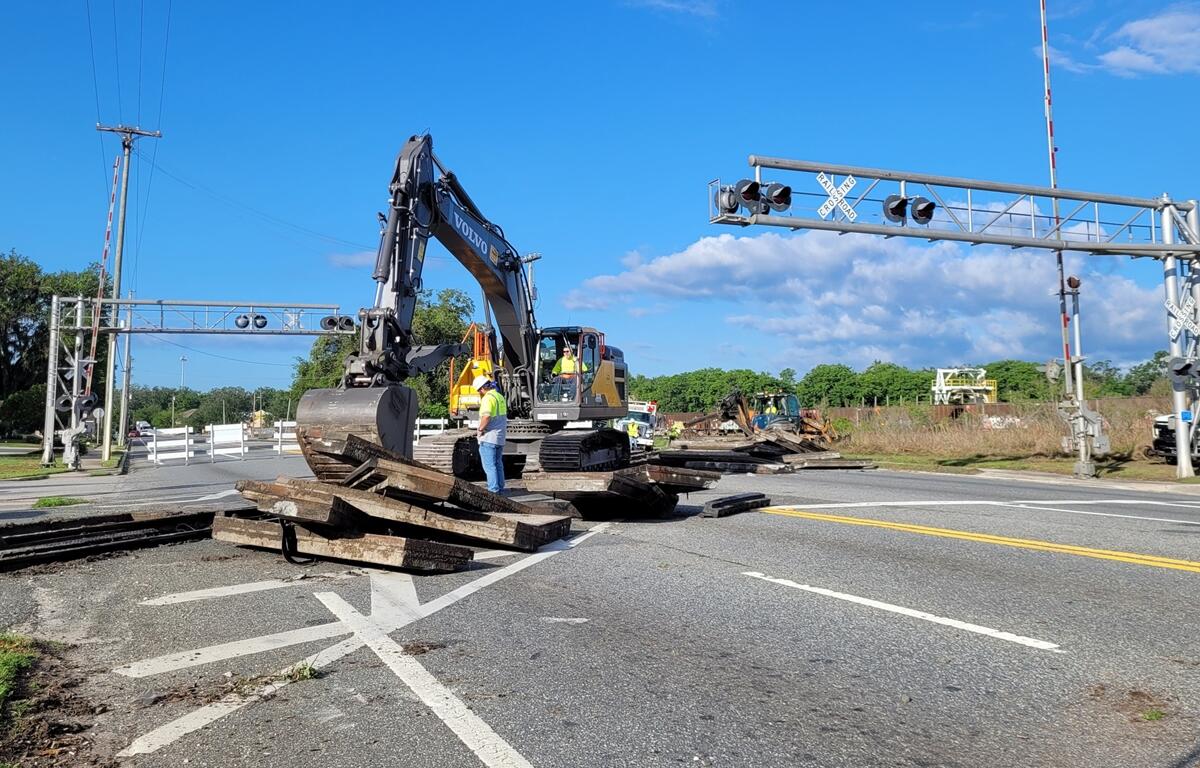 Construction workers use heavy equipment to remove wooden railroad ties from a crossing on U.S. 301 in Wildwood, Florida, with traffic signals and warning gates visible above the road.