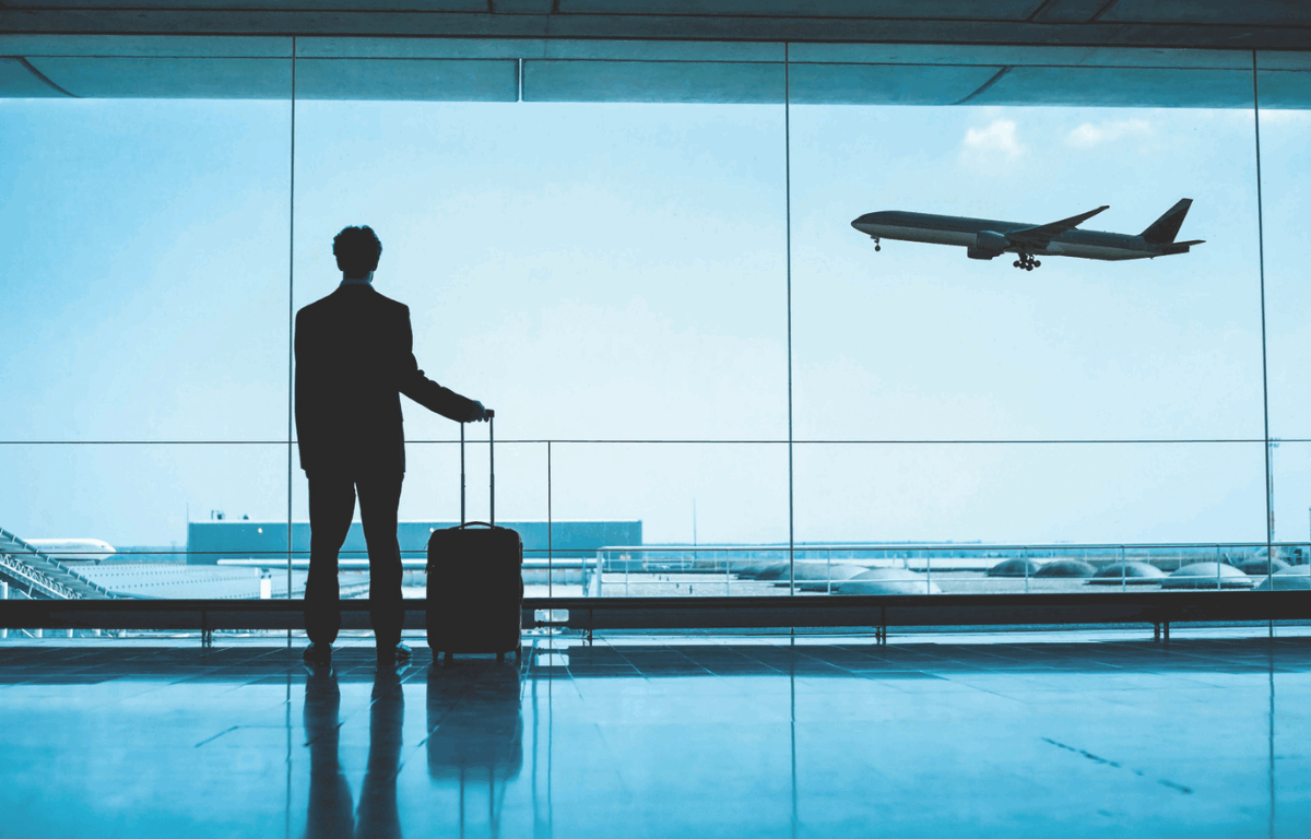 A silhouette of a man in a suit stands in an airport terminal with a rolling suitcase, gazing out a large window as a commercial airplane takes off into the sky.