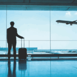 A silhouette of a man in a suit stands in an airport terminal with a rolling suitcase, gazing out a large window as a commercial airplane takes off into the sky.