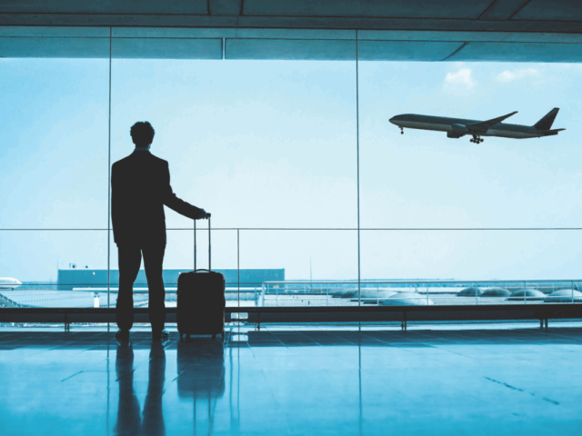 A silhouette of a man in a suit stands in an airport terminal with a rolling suitcase, gazing out a large window as a commercial airplane takes off into the sky.