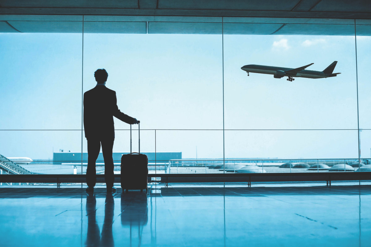 A silhouette of a man in a suit stands in an airport terminal with a rolling suitcase, gazing out a large window as a commercial airplane takes off into the sky.