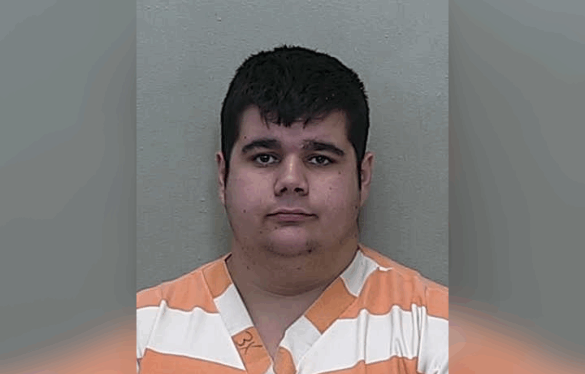 Mugshot of a young adult male with short dark hair wearing an orange and white striped prison uniform, standing against a neutral gray background.