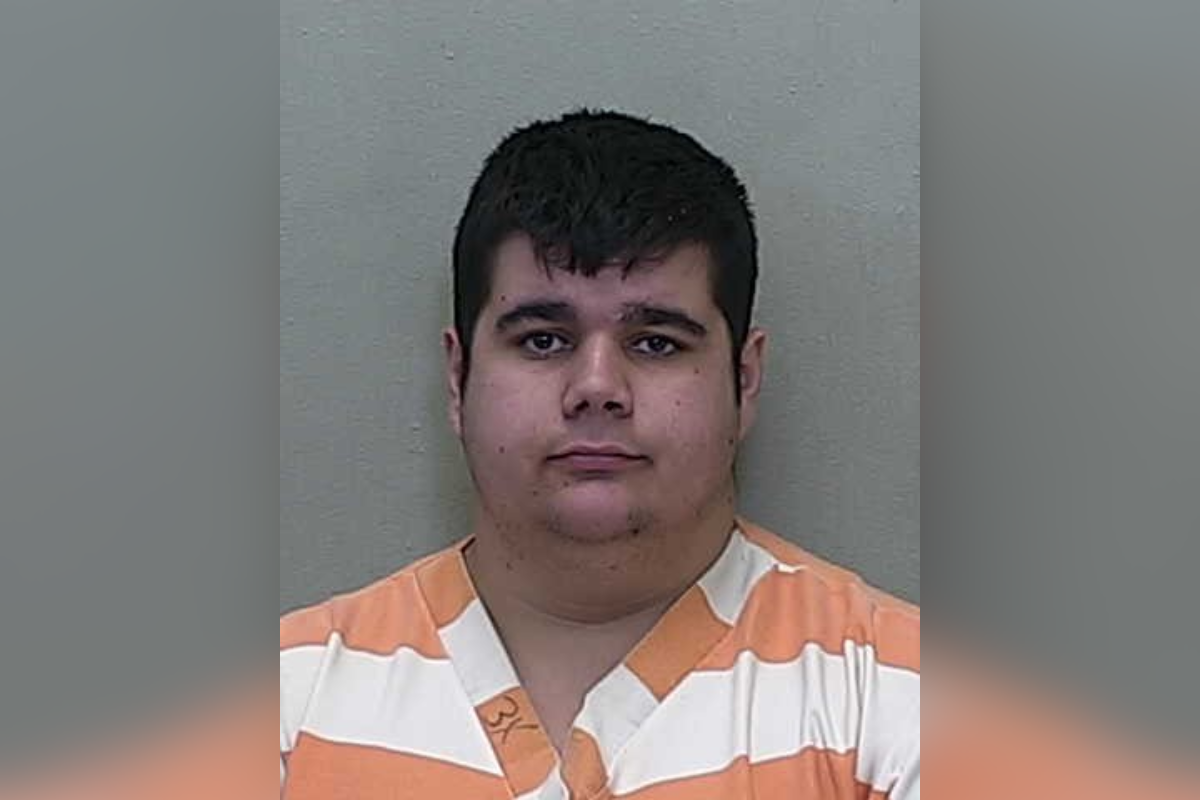 Mugshot of a young adult male with short dark hair wearing an orange and white striped prison uniform, standing against a neutral gray background.