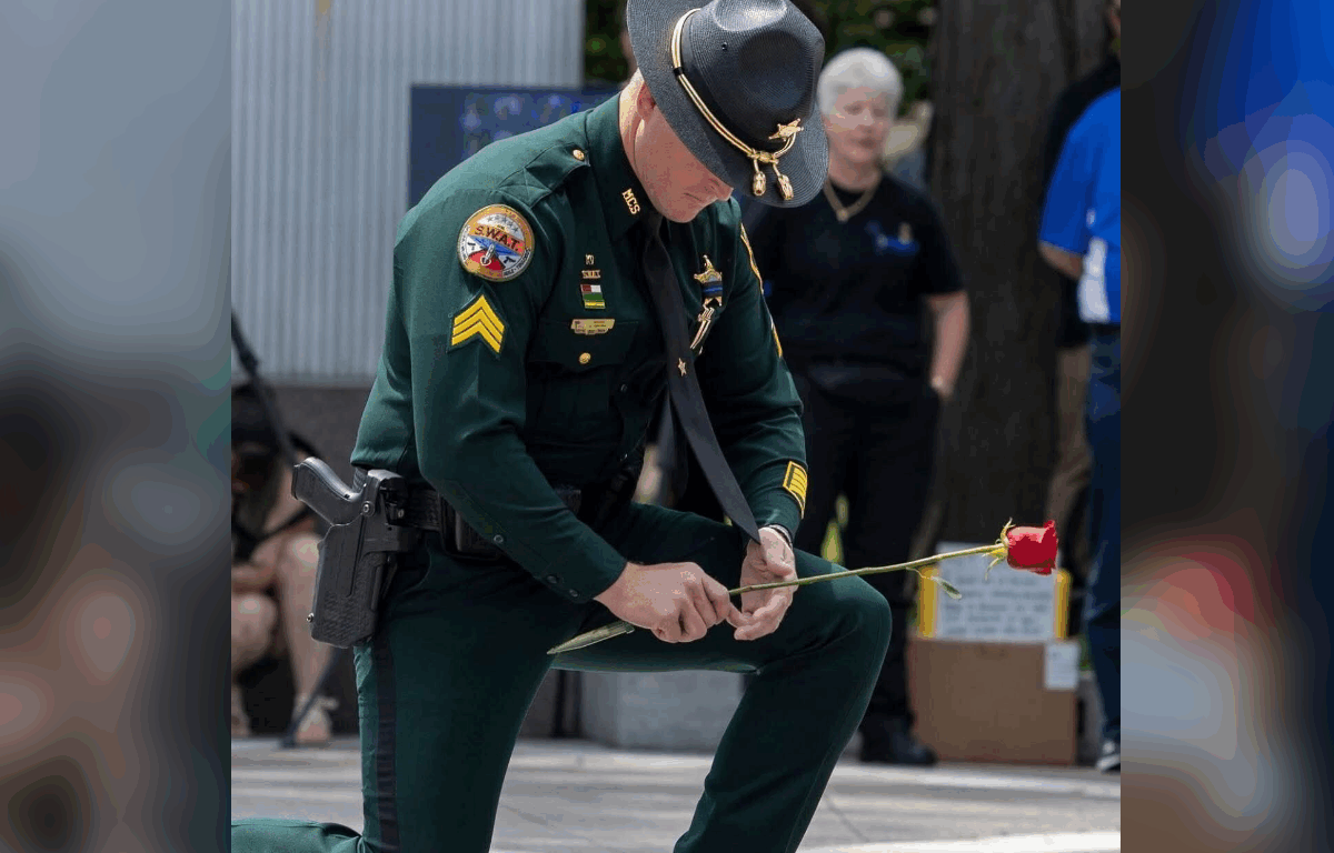 A uniformed law enforcement officer kneels in solemn tribute while holding a single red rose during a memorial ceremony, honoring fallen officers.