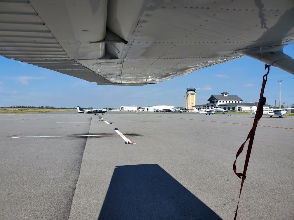 View from underneath the wing of a parked aircraft on a sunny day at a small airport. Several light aircraft are lined up on the tarmac, with a control tower and terminal building visible in the background. A red tie-down strap hangs from the wing in the foreground, casting a shadow on the pavement. The sky is mostly clear with a few clouds.