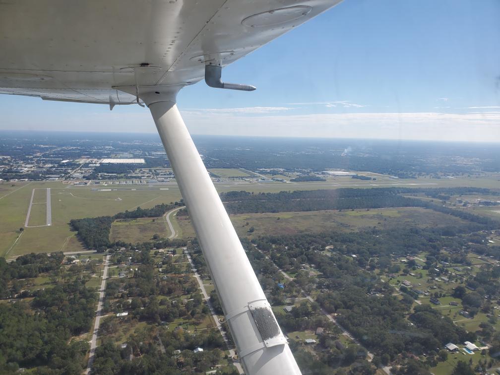 Aerial view from inside a small aircraft looking out over a city and surrounding landscape. The aircraft’s wing and strut are visible in the foreground, with a runway and airport facilities seen below. Residential neighborhoods, roads, and patches of forest spread out across the landscape under a clear blue sky.