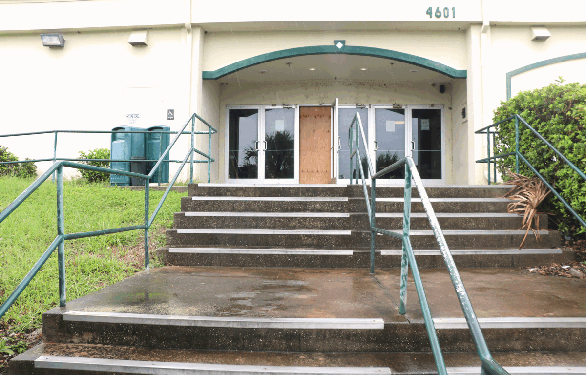 A set of wet concrete steps leads to the entrance of a building at 4601, with metal railings and a row of glass doors, one of which is boarded up with plywood. Rain is visible on the ground and handrails, and the surrounding grass appears damp and slightly overgrown.