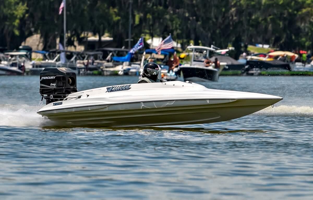 A white drag boat with a Mercury engine races on Lake Eustis with spectators watching from boats in the background.