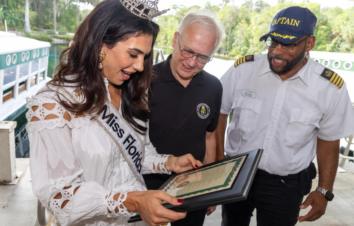 Miss Florida holding a certificate while standing with two men near the water.
