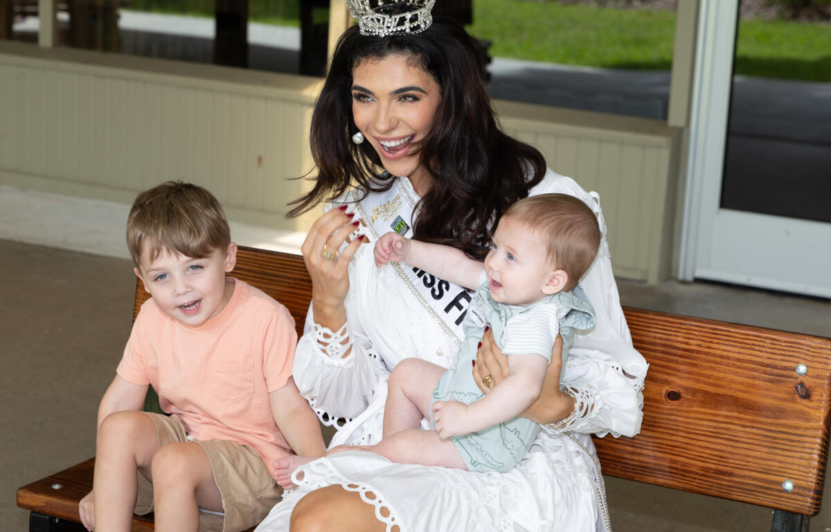 Miss Florida sitting on a bench holding a baby and smiling with a toddler beside her.