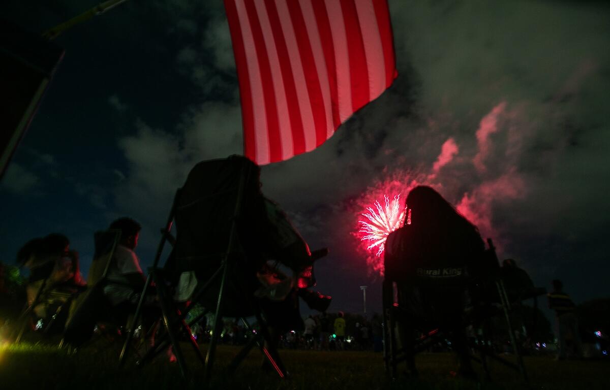 People sitting in lawn chairs watch fireworks under an American flag at night during a Fourth of July event