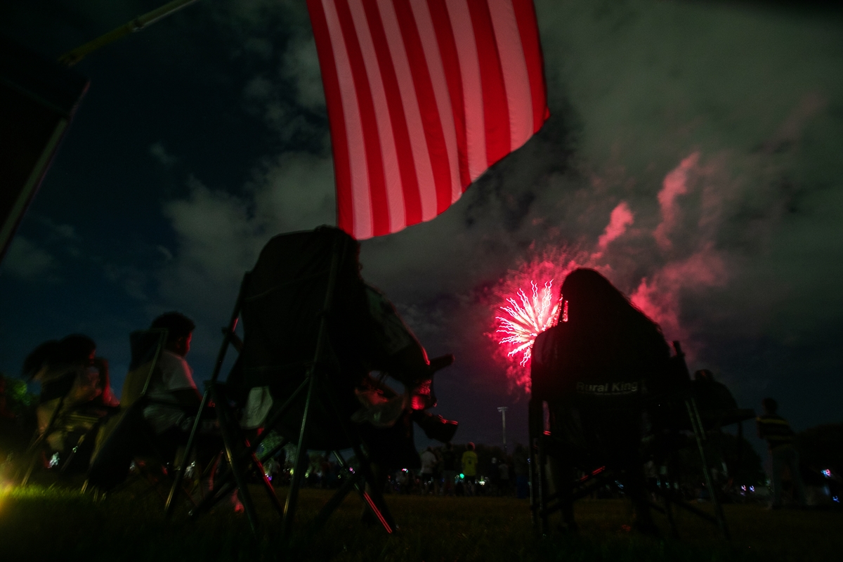 People sitting in lawn chairs watch fireworks under an American flag at night during a Fourth of July event