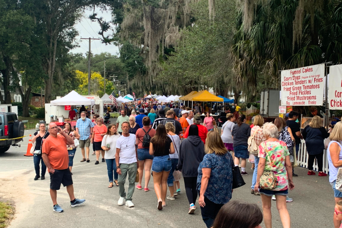 Crowds of people walk through a lively outdoor festival lined with vendor tents and food stands.