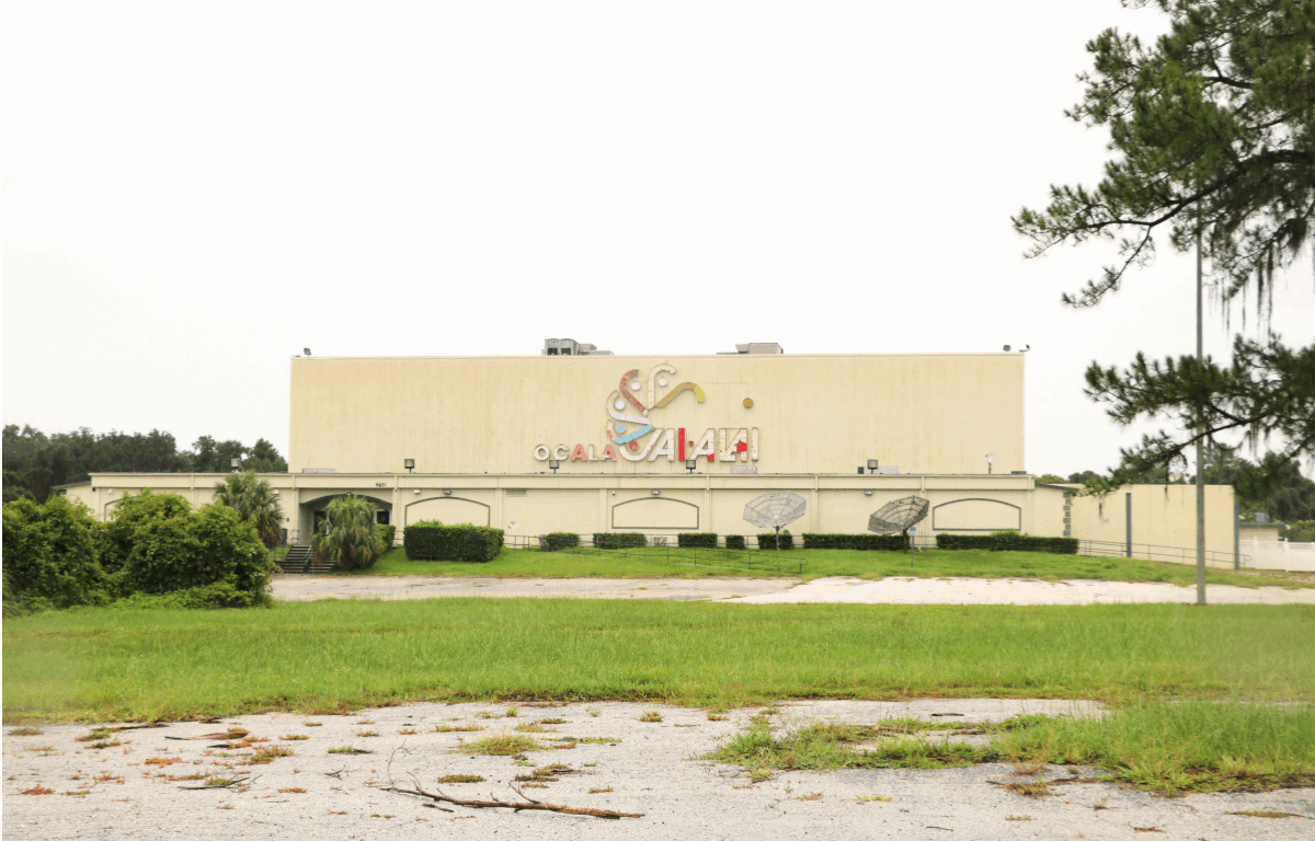 A wide view of the Ocala Jai-Alai building shows a large beige structure with colorful signage and satellite dishes on the lawn. The front area is overgrown with patches of grass and scattered debris, suggesting the site is no longer in active use.