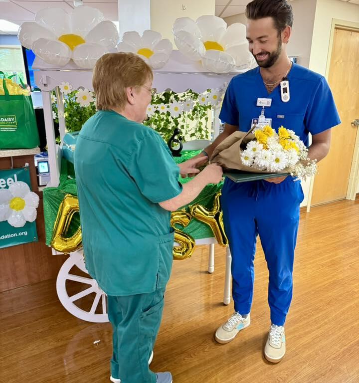 Dustten Martineau smiles while receiving flowers from a colleague in front of a daisy-themed DAISY Award display cart.