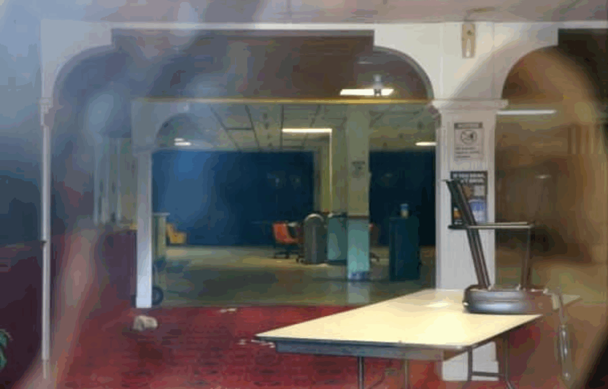 Interior view of a dimly lit, abandoned or vacant building with arched white pillars, red patterned carpet, and scattered furniture. A folding table with an upside-down chair sits in the foreground, while a row of orange chairs and items are visible deeper inside the building. The scene has a slightly, eerie, empty atmosphere.