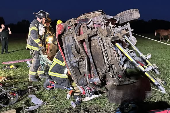 Several firefighters in full gear work at the scene of a serious vehicle accident involving a red SUV that has flipped onto its side. The vehicle is heavily damaged and propped up with stabilization tools. Debris and rescue equipment are scattered around the grassy field as emergency responders assess the situation in low light.
