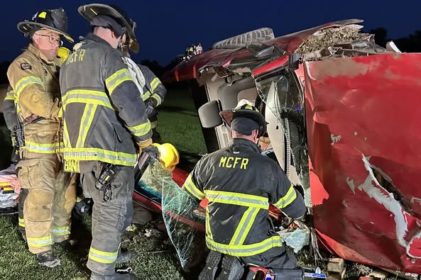 Firefighters from MCFR work to extract an individual from the overturned red SUV. The vehicle's side is crushed, and broken glass is visible as the responders use specialized tools to access the interior. The scene is lit by portable lights during nighttime rescue operations.