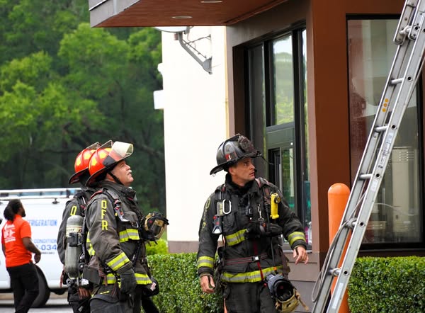 Three Ocala Fire Rescue firefighters in full gear look up toward the roof of a Dunkin' Donuts building beside a ladder. One firefighter holds a tool while the others observe attentively.