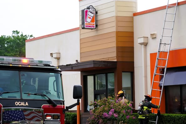 Firefighters stand near a ladder propped against the side of a Dunkin' Donuts building in Ocala, Florida, with a fire truck parked out front. One firefighter is climbing the ladder while another stands nearby among bushes.