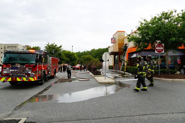 Multiple fire trucks and firefighters are on scene at a Dunkin' Donuts parking lot, with hoses on the wet pavement and a “Do Not Enter” sign near the building. The emergency response appears organized and active.