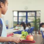 a young girl in a school cafeteria holding a lunch tray. On the tray, there's an apple, a wrap or burrito, some leafy greens, and a green drink container.