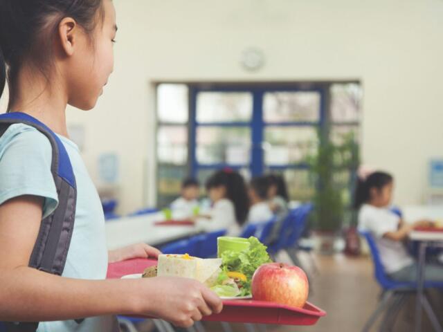 a young girl in a school cafeteria holding a lunch tray. On the tray, there's an apple, a wrap or burrito, some leafy greens, and a green drink container.