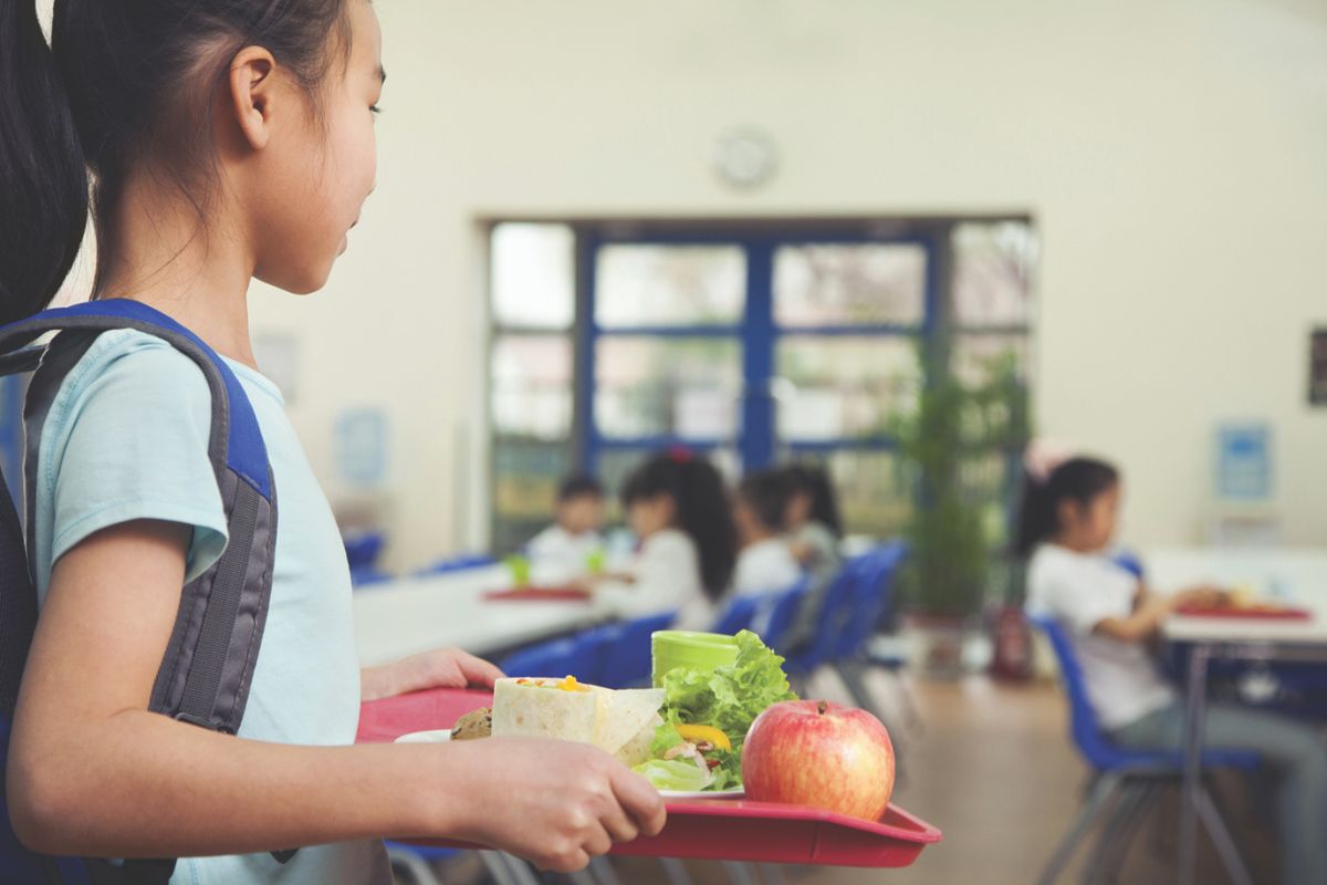 a young girl in a school cafeteria holding a lunch tray. On the tray, there's an apple, a wrap or burrito, some leafy greens, and a green drink container.