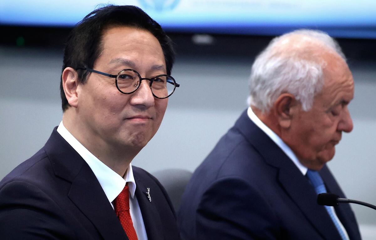Santa Ono sits beside another official during a meeting, wearing a dark suit, red tie, and glasses, as he looks toward the camera.