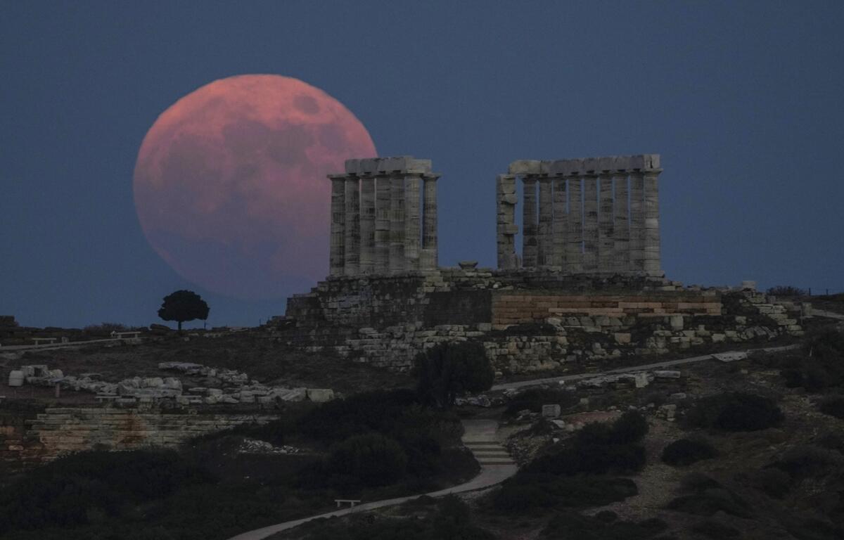 A large reddish full moon rises behind the ruins of an ancient stone structure on a hill, silhouetted against a deep blue evening sky.