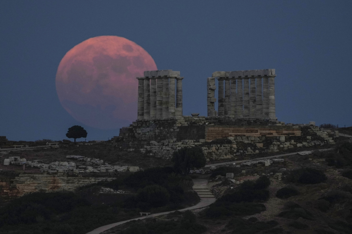 A large reddish full moon rises behind the ruins of an ancient stone structure on a hill, silhouetted against a deep blue evening sky.