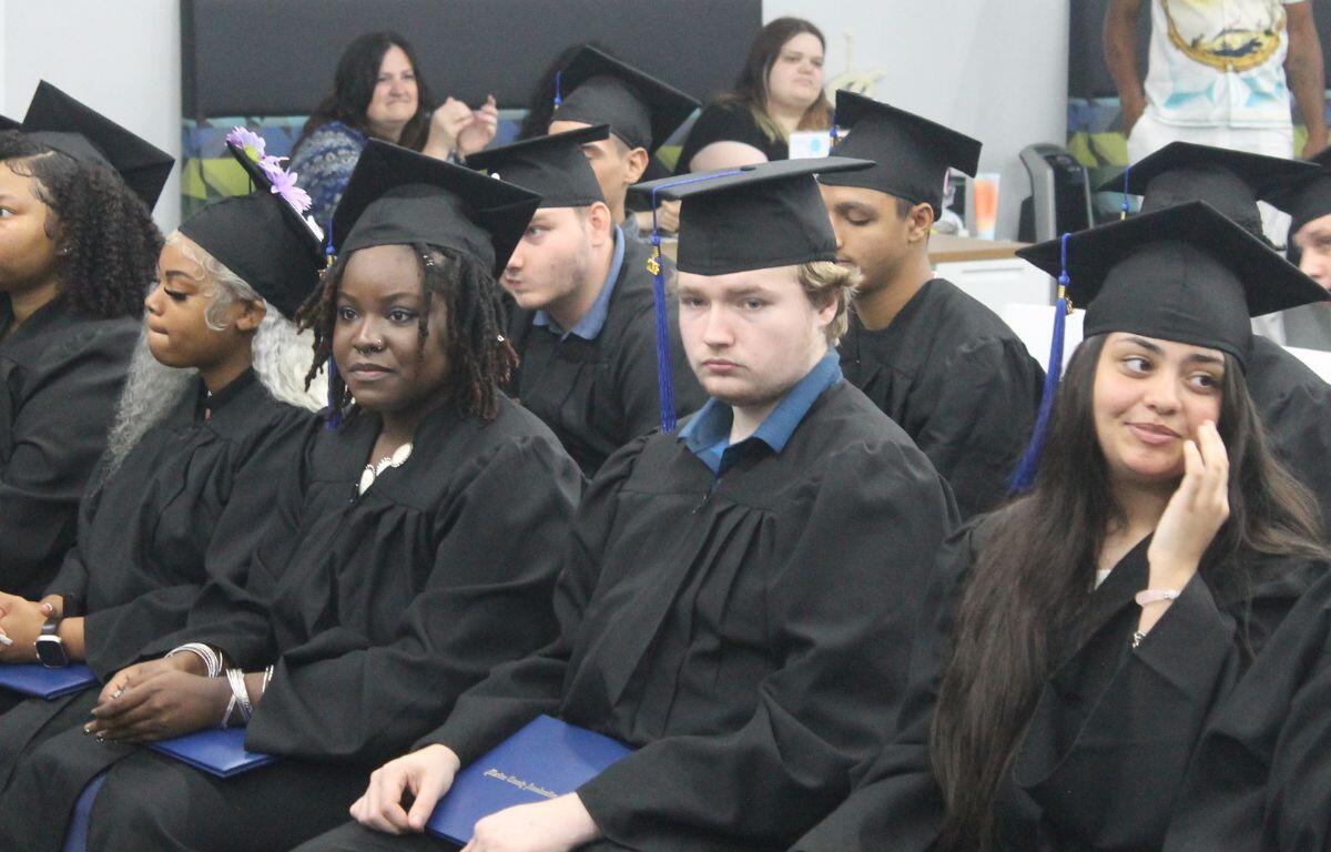 Students sitting side by side in their cap and gown and holding their diplomas, with other students behind them, and staff members sitting against the wall in the background.