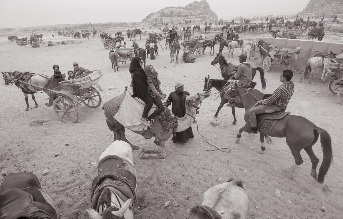 People on camels, horses, and carriages gather near ancient ruins on the sandy terrain of Giza, Egypt, in a busy tourist area.
