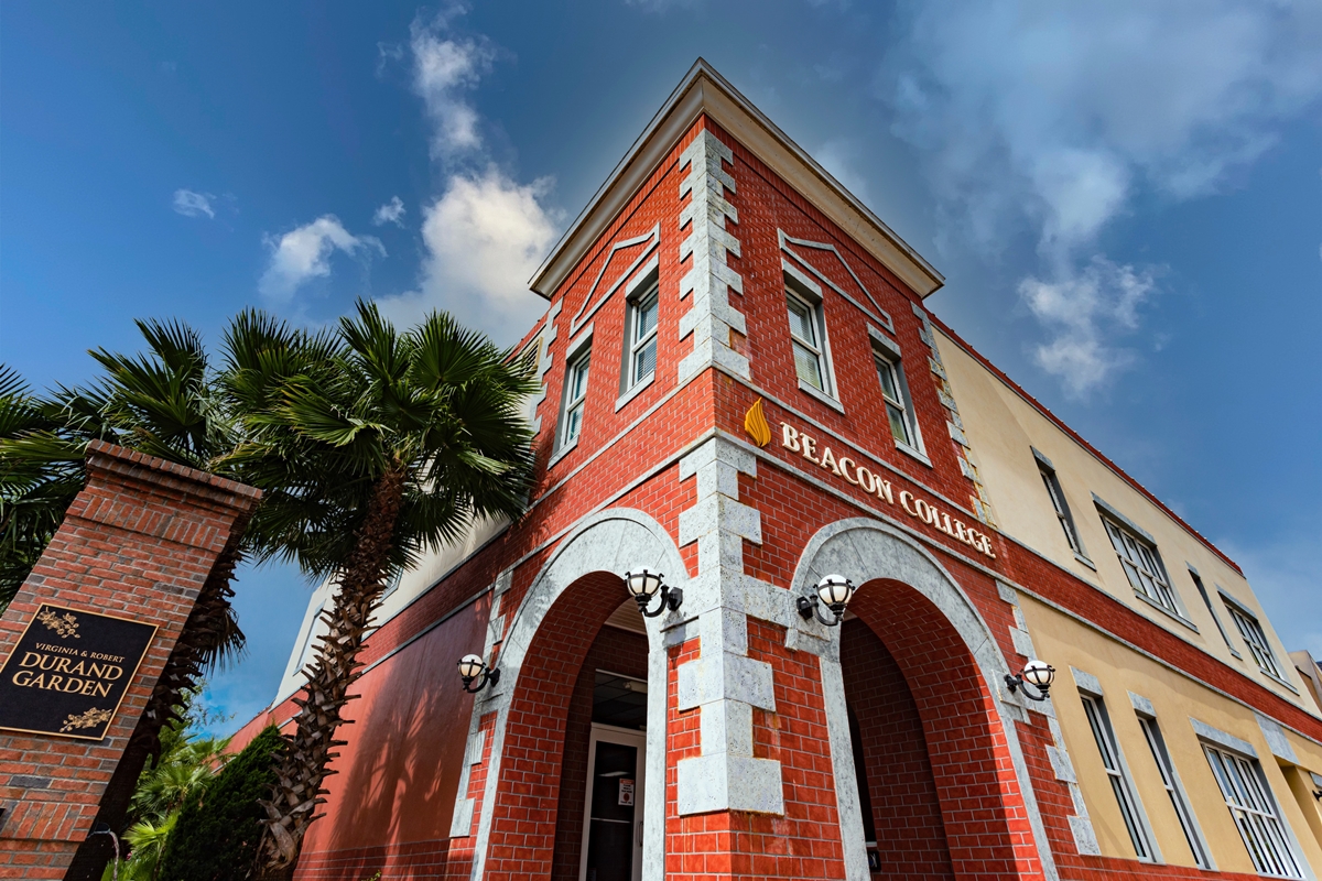Red brick exterior of Beacon College building in Leesburg, Florida, with arched entryway and palm trees nearby.