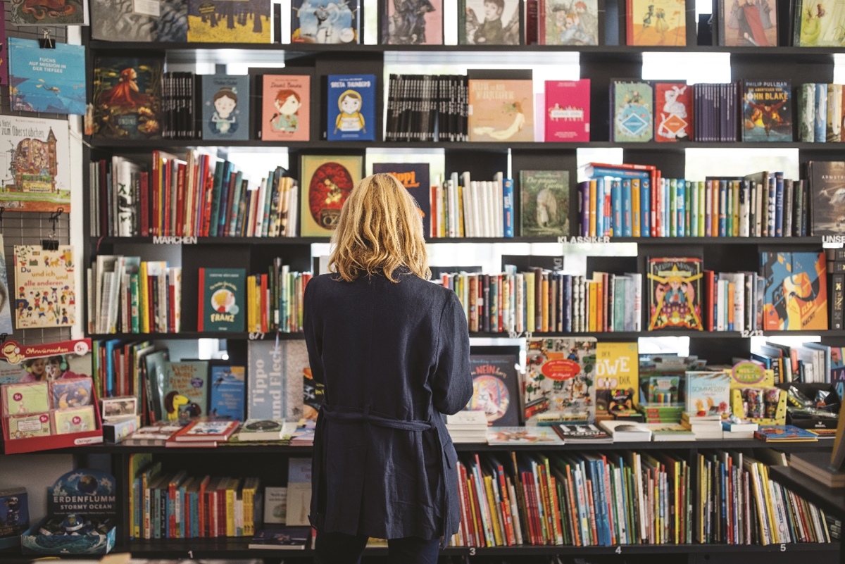 Person browsing shelves of books at a library sale with various children's and illustrated books on display.