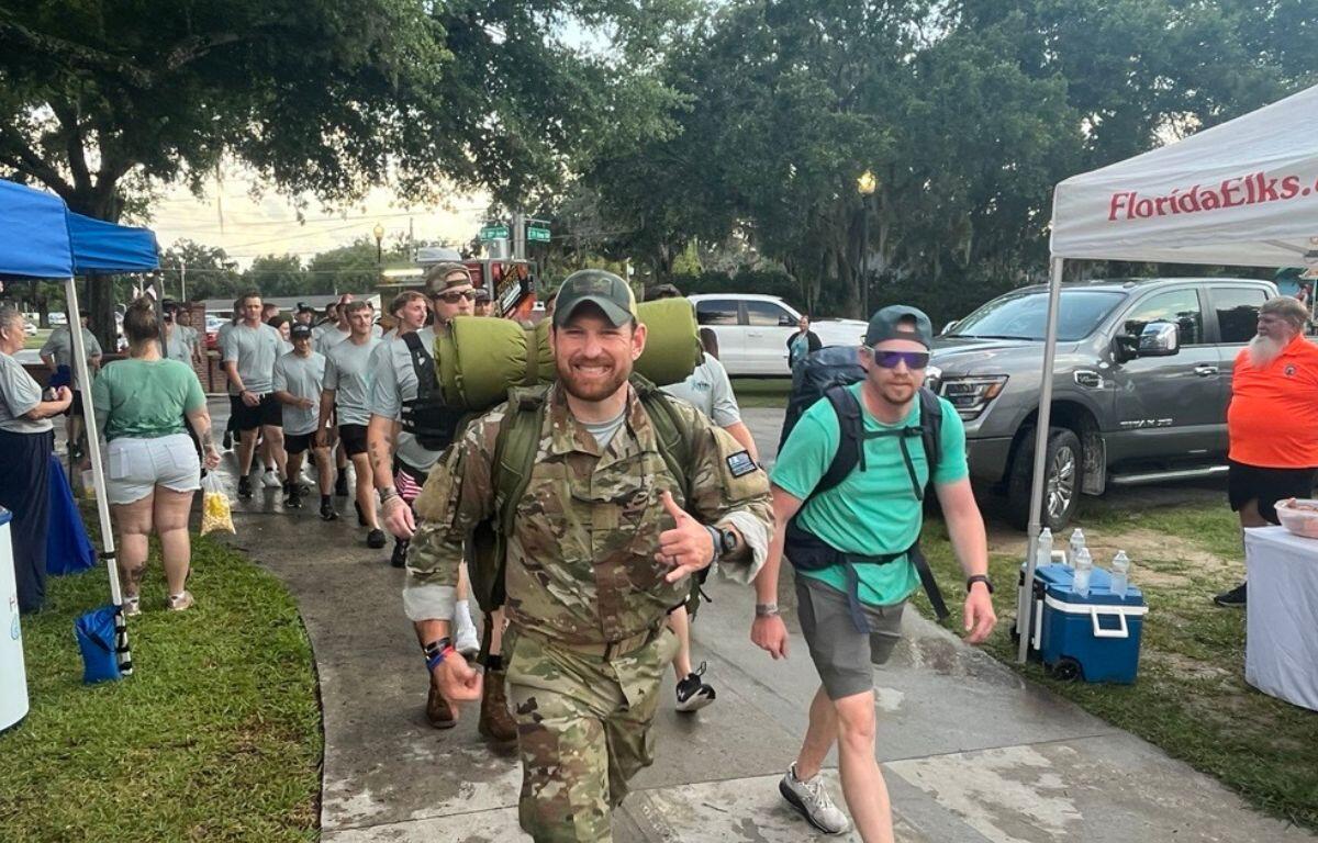 Grass to the left, two men walking on a gray concrete pathway, the one on the left dressed in combat fatigues, the man on the right in a green shirt, both wearing backpacks, with vehicles and tents to their right, and with people in the background, walking under a canopy of trees.